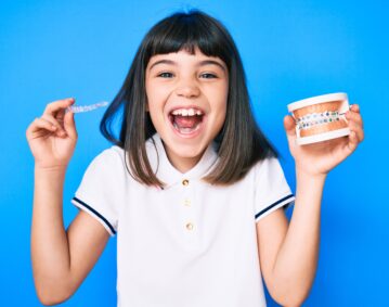 Young little girl with bang holding invisible aligner orthodontic and braces smiling and laughing hard out loud because funny crazy joke.